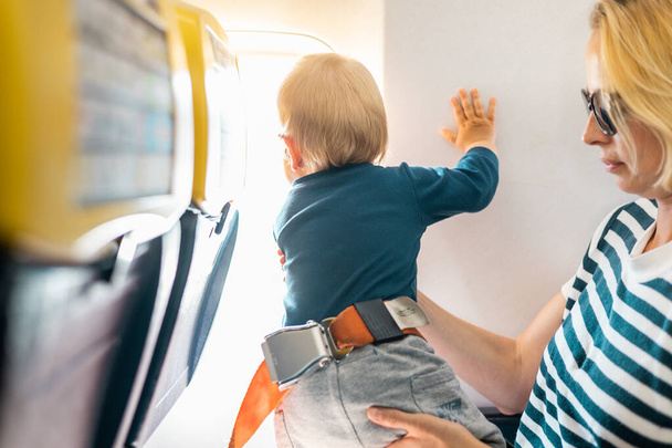 Äiti ja lapsi lentävät lentokoneella. Äiti pitelee ja leikkii pikkupoikalapsensa kanssa tämän sylissä turistiluokan lennon aikana. Konsepti kuva lentomatkustamisesta vauvan kanssa. Oikeat ihmiset - Valokuva, kuva