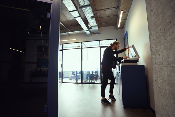 Bearded male in casual clothes uses a photocopier in a coworking space, the room is clean and spacious - Photo, Image