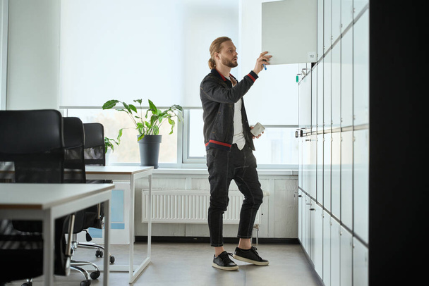 Young man opens the door of a locker in a coworking space, the male has a cup in his hands - Foto, afbeelding