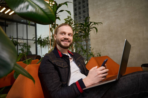 Young bearded man sits in a lounge area with a mobile phone and a laptop, living plants around - Photo, image