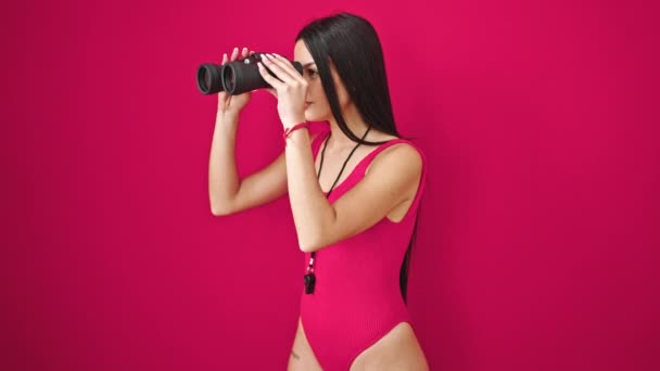 Joven bella salvavidas hispana usando prismáticos con expresión sorpresa sobre fondo rojo aislado - Imágenes, Vídeo