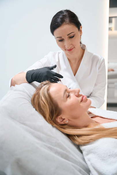 Beautician in uniform performs an injection procedure around the eyes of a blonde woman, an esthetician uses a thin needle - Foto, imagen