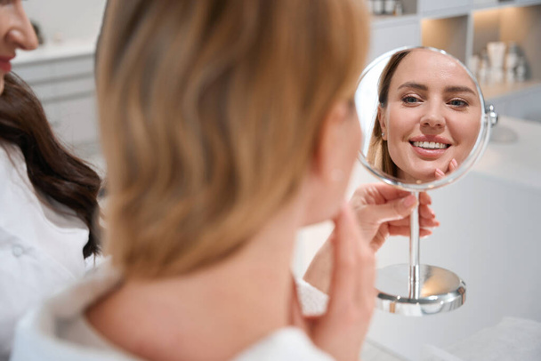 Glückliche Blondine blickt bei einer Beratung mit einem Schönheitschirurgen in den Spiegel, sie freut sich über die Spiegelung im Spiegel - Foto, Bild