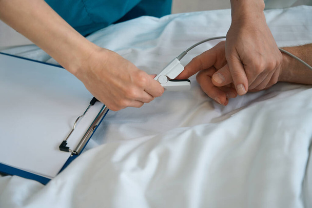 Zbliżenie lekarza umieszczającego oksymetr pulsacyjny na męskim palcu pacjenta, który leży w łóżku w klinice - Zdjęcie, obraz