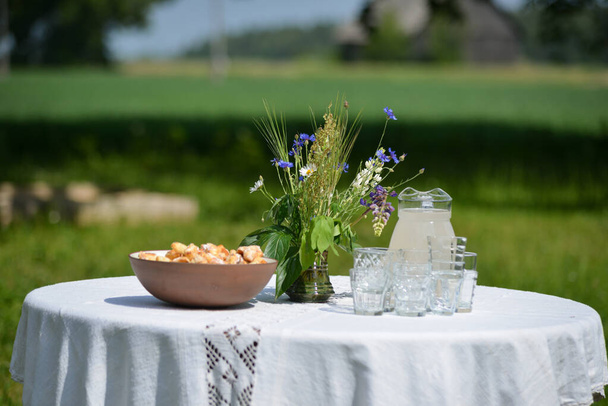 Mesa de celebração de verão com toalha de mesa branca. sobre os copos de cristal de mesa com tortas em uma tigela de barro marrom e flores de verão em uma caneca de barro em um dia quente de verão com um fundo borrado verde. - Foto, Imagem