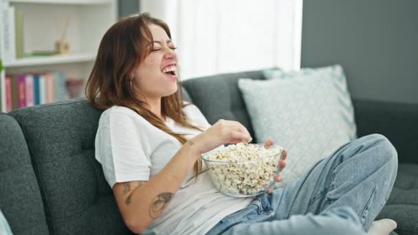 Mladá krásná hispánská žena sleduje komediální film jíst popcorn sedí na pohovce doma - Záběry, video