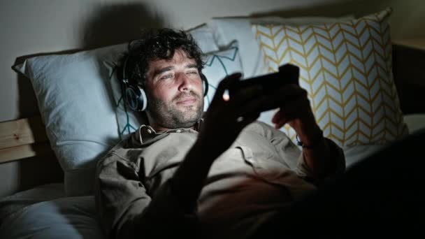 Молодий іспаномовний чоловік грає в відеогру, лежачи на ліжку в спальні - Кадри, відео