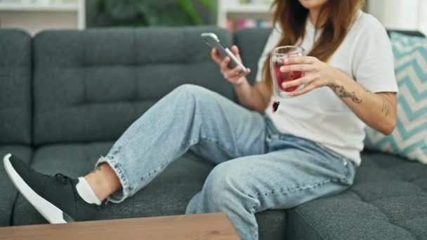 Νεαρή όμορφη ισπανόφωνη γυναίκα που χρησιμοποιεί smartphone ξαπλωμένη στον καναπέ κρατώντας ένα ποτήρι τσάι στο σπίτι - Πλάνα, βίντεο