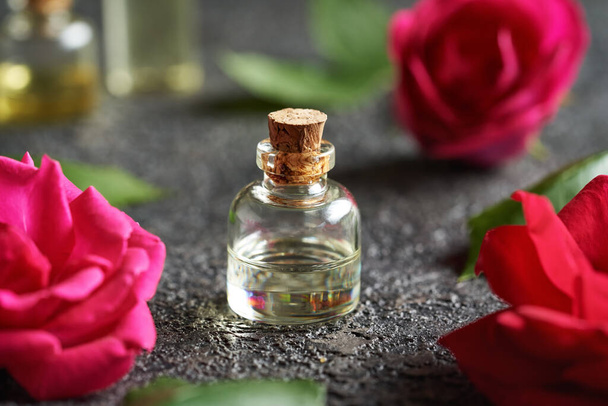 A bottle of essential oil with fresh red rose flowers and leaves on dark background - Фото, зображення