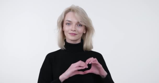 Beautiful Caucasian woman stands against a clean white background, radiating warmth and affection. With a genuine smile and a sparkle in her eyes, she lovingly forms a heart shape with her hands. - Footage, Video