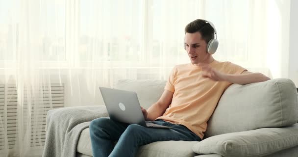 Кавказький чоловік сидить на дивані, носить навушники і захоплюється відеодзвінком на свій ноутбук. З орієнтованим виразом він використовує сучасні технології для зв'язку з іншими у віртуальному світі. - Кадри, відео