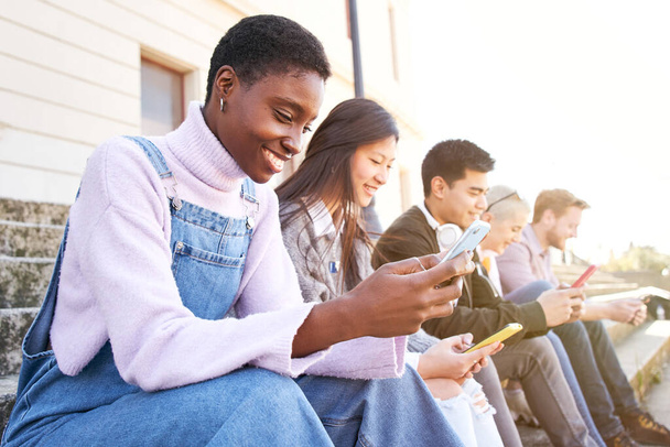 Technologia Uzależniona uśmiechnięta grupa radosnych uczniów korzystających z telefonów komórkowych. Sprawdzają multimedia w swoich telefonach szczęśliwy i nie patrzą na siebie. Śmieją się na dworze. - Zdjęcie, obraz