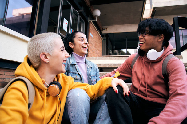 Group of three students laugh a lot sitting outdoors. Teenage gen z multiracial classmates are happy and cheerful. Young colleagues at high school having fun together. Friendship and community - Photo, Image