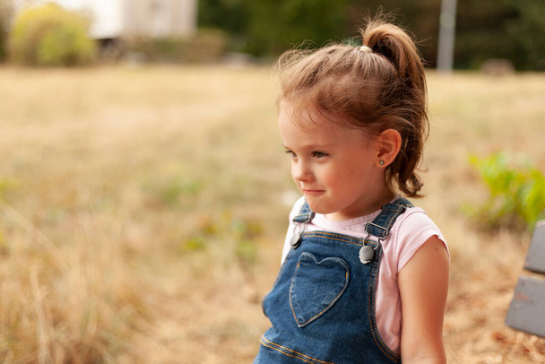 A beautiful blonde little girl with a tail sits on a bench in the park in a denim overalls - Fotó, kép