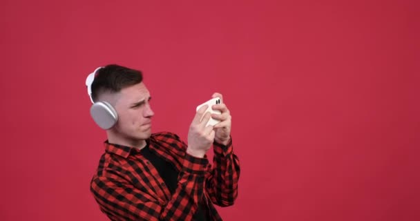 Na červeném pozadí, běloch ve sluchátkách je zaujat hraním hry na svém telefonu. Se soustředěnou pozorností drží sluchátko v rukou, klepe na obrazovku a šmátrá po ní.. - Záběry, video
