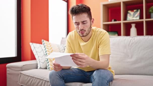 Young hispanic man reading document sitting on the sofa at home - Footage, Video