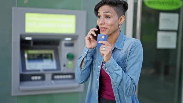 銀行テラーでクレジットカードを保持しているスマートフォンで話す若い美しいヒスパニックの女性 - 映像、動画