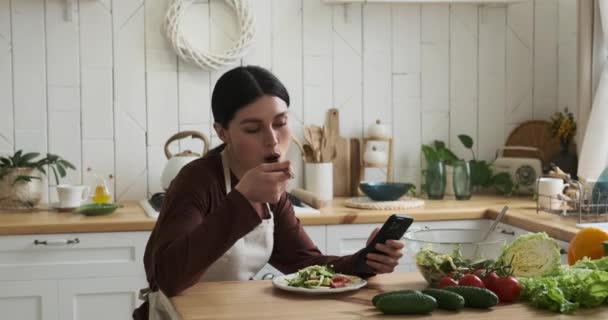 Kaukasische Frau von charmanter Anmut sitzt an einem Küchentisch und genießt einen köstlichen Salat von ihrem Teller. Sie hält das Telefon in der anderen Hand, blickt gelegentlich auf den Bildschirm und teilt freudiges Lachen.. - Filmmaterial, Video