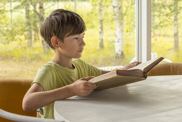 Ребенок садится за стол и читает старую книгу. Мальчик с книгой в руке. Детский урок с книгой - Фото, изображение