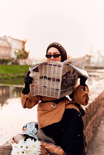 Kobieta w hidżabie z wdziękiem ozdobiona fuzją starych i nowoczesnych francuskich strojów, zanurzona w tętniących życiem ulicach miasta, pochłonięta czytaniem gazety.  - Zdjęcie, obraz