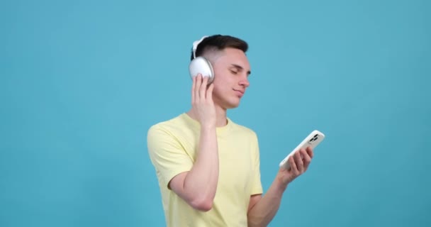 青い背景には、陽気な白人男性が携帯電話を手に音楽を楽しんでいるのが見えます。耳を満たすメロディーに身を浸しながら、明るい笑顔が彼の顔を飾ります. - 映像、動画