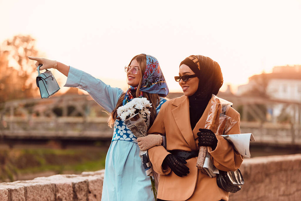 ヒジャーブと現代的で伝統的なドレスを着たカップルの女性と、青いドレスとスカーフを着たカップルは、日没時に街を歩いています。花束とパンを持っているのに対し - 写真・画像