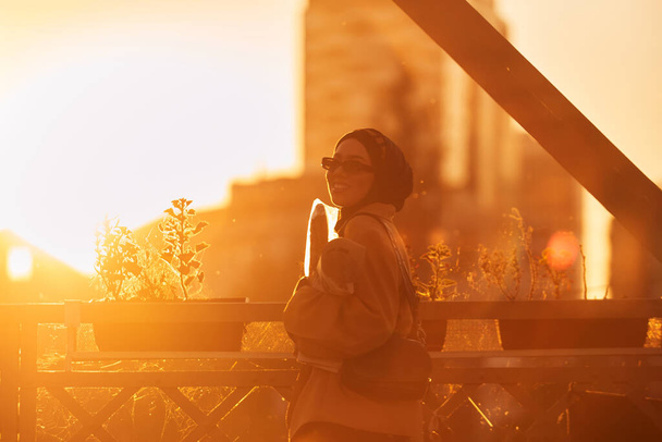 Eine Hijab-Frau mit stylischer Sonnenbrille und elegantem französischen Outfit spaziert bei Sonnenuntergang durch die Stadt, trägt einen Strauß, Brot und Zeitungen bei sich und strahlt einen Sinn für kulturellen Charme und Gelassenheit aus..  - Foto, Bild