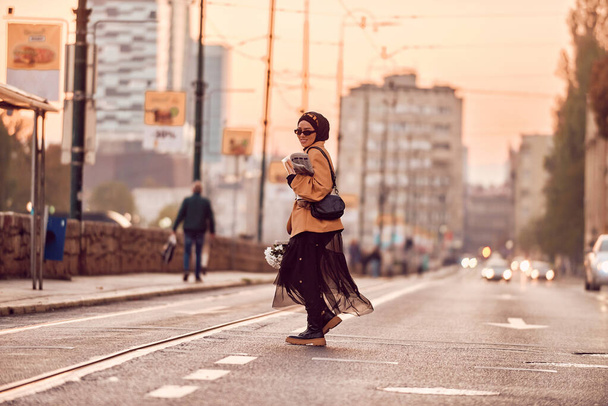 Μια γυναίκα hijab, φορώντας ένα πολυτελές αντίκα γαλλική στολή, καθώς περπατά στους δρόμους της πόλης στο ηλιοβασίλεμα, μεταφέρουν ψωμί, ένα μπουκέτο λουλούδια, και εφημερίδες στο χέρι της, προκαλώντας ένα σαγηνευτικό μείγμα - Φωτογραφία, εικόνα