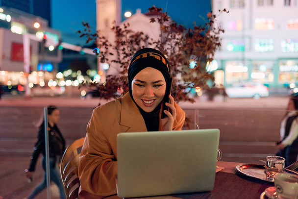 Una mujer hijab que usa un ordenador portátil y un teléfono inteligente en un café moderno en el paisaje urbano nocturno, lo que refleja la armoniosa mezcla de tecnología y ambiente urbano.  - Foto, imagen