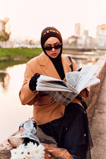 Una mujer en un hiyab elegantemente adornada con una fusión de atuendo francés antiguo y moderno, inmersa en las bulliciosas calles de la ciudad, absorta en la lectura de un periódico.  - Foto, imagen