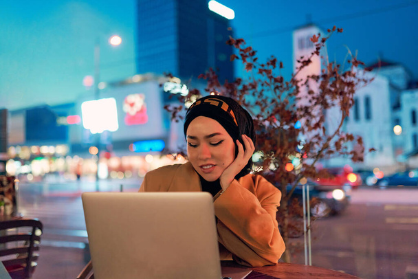 En el encantador ambiente de un paisaje urbano nocturno, una chica vestida de hiyab absorta en su computadora portátil crea una imagen fascinante, que encarna la fusión de la tecnología, el empoderamiento y la vibrante ciudad. - Foto, Imagen
