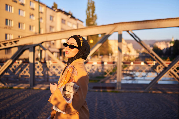 Hidżabska kobieta w stylowych okularach przeciwsłonecznych i eleganckim francuskim stroju, przechadzająca się po mieście o zachodzie słońca, niosąca bukiet, chleb i gazetę, promieniująca poczuciem kulturowego uroku i spokoju.  - Zdjęcie, obraz