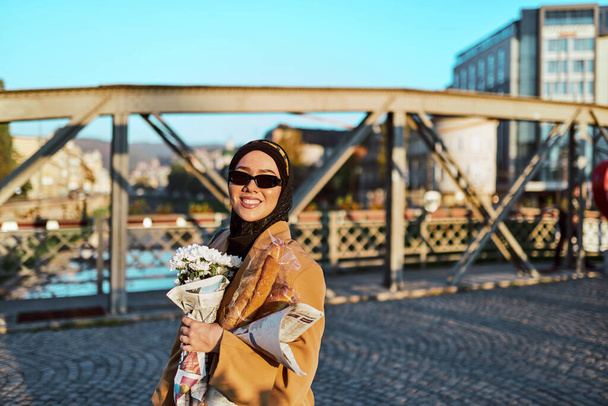 A hijab woman in stylish sunglasses and an elegant French outfit, walking through the city at sunset, carrying a bouquet, bread, and newspaper, radiating a sense of cultural charm and serenity.  - Фото, изображение