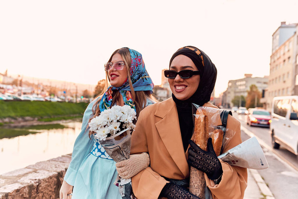 Couple woman one wearing a hijab and a modern yet traditional dress, and the other in a blue dress and scarf, walking together through the city at sunset. One carries a bouquet and bread, while the - Фото, изображение