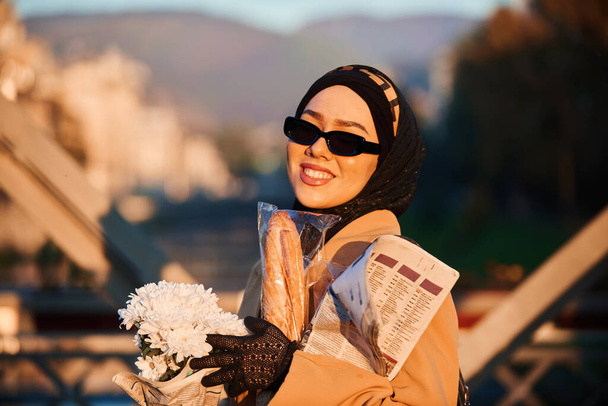 スタイリッシュなサングラスとエレガントなフランスの服を着たヒジャーブの女性は、日没時に街を歩き、花束、パン、新聞を運び、文化的な魅力と静けさの感覚を放射します.  - 写真・画像