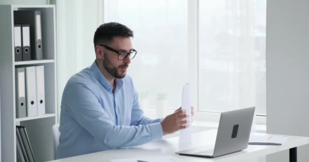 Blanke zakenman met een baard en bril glimlacht terwijl hij een videogesprek voert via zijn laptop. Hij demonstreert documenten met grafieken en groeistatistieken. - Video