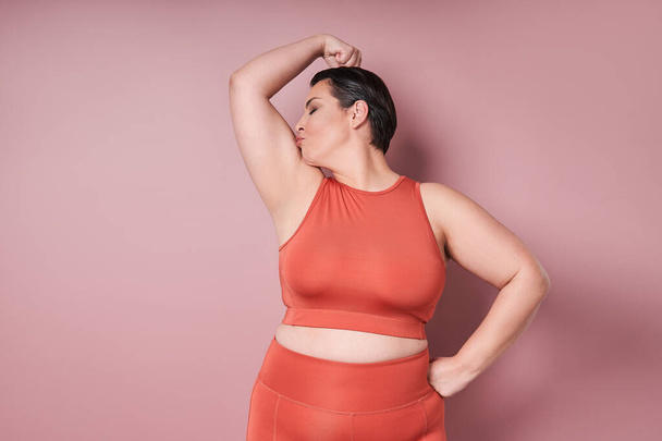 Young caucasian plus size female model kissing biceps after training on pink background. Concept of sport, healthy lifestyle, body positive, equality - Foto, imagen