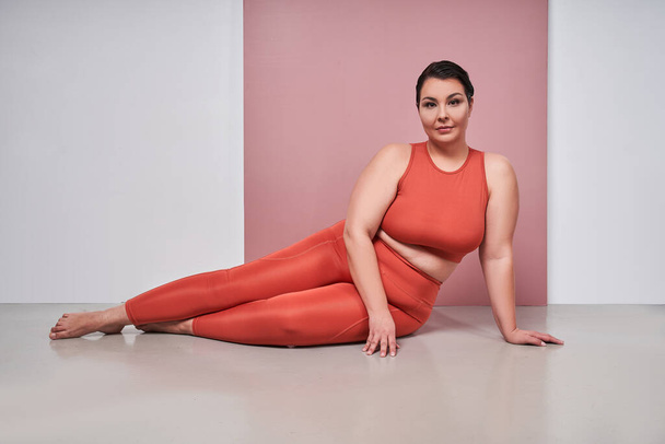 Smiling overweight woman in sportswear isolated on pink studio background feel body positive. Happy fat girl show natural shape and curvy figure - Foto, Bild
