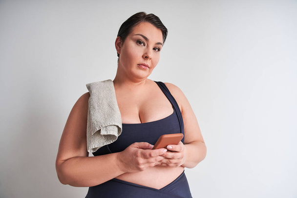 Sporty fat woman holding smartphone and poses camera in white wall studio background. Healthy lifestyle and people concept - Fotó, kép