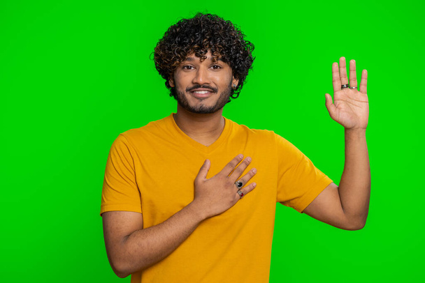 Ich schwöre, ehrlich zu sein. Aufrichtiger, verantwortungsvoller junger indischer Mann, der die Hand zum Eid erhebt und verspricht, ehrlich zu sein und die Wahrheit zu sagen und die Hand auf der Brust zu halten. Hindu-Kerl isoliert auf grünem Chroma-Schlüsselhintergrund - Foto, Bild