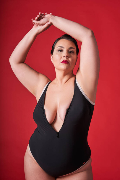 Feeling proud in the studio. Satisfied woman standing against red studio background. Body positive woman feeling comfortable in underwear - Photo, Image