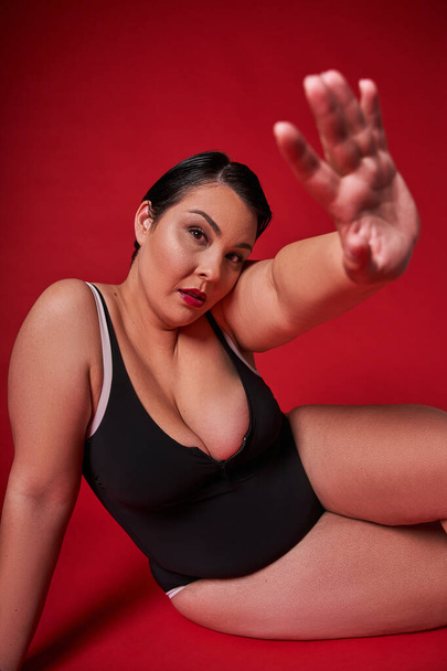 Plus size self enjoyed woman wearing lingerie sitting on the floor isolated over red background. Body positive concept - Foto, Imagem