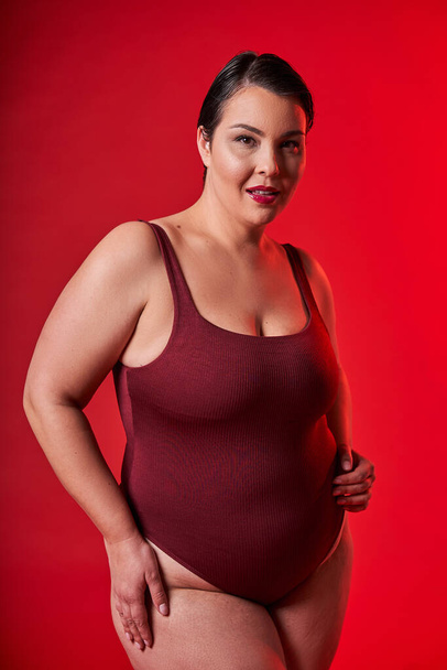 Feeling happy in the studio. Smiling woman standing against red studio background. Body positive woman feeling comfortable in underwear - Foto, afbeelding