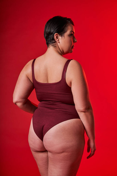 Stop body shaming concept. Curvy asian woman in underwear standing over red studio background, looking aside at free space, back view - 写真・画像