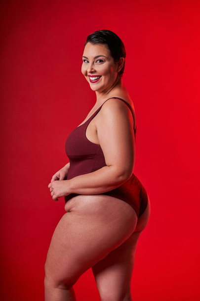 Plus size model. Profile portrait of half-turned smiling lady in underwear standing over red studio background, looking at camera. Female in lingerie posing - Foto, Bild