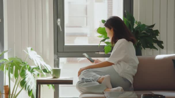 Asian brunette girl scrolling feed at her smartphone and preparing drinking hot beverage near the window at the morning. Drinks and people concept - Materiaali, video