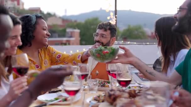 Diverse happy friends having dinner party outside sitting around table at rooftop terrace. Millennial people enjoying a meal during sunset, celebrating together, laughing and having fun. - Footage, Video
