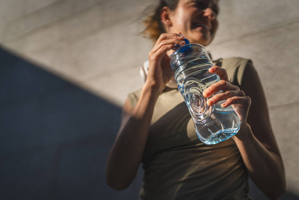 primer plano en las manos sección media de mujer caucásica desconocida abierta botella de plástico de agua, mientras que de pie al aire libre en el día soleado - Foto, imagen
