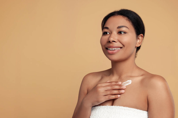 Young woman applying cream onto body on beige background. Space for text - Foto, afbeelding