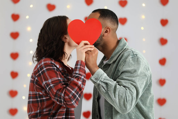 Lovely couple kissing behind red paper heart indoors. Valentine's day celebration - Fotografie, Obrázek